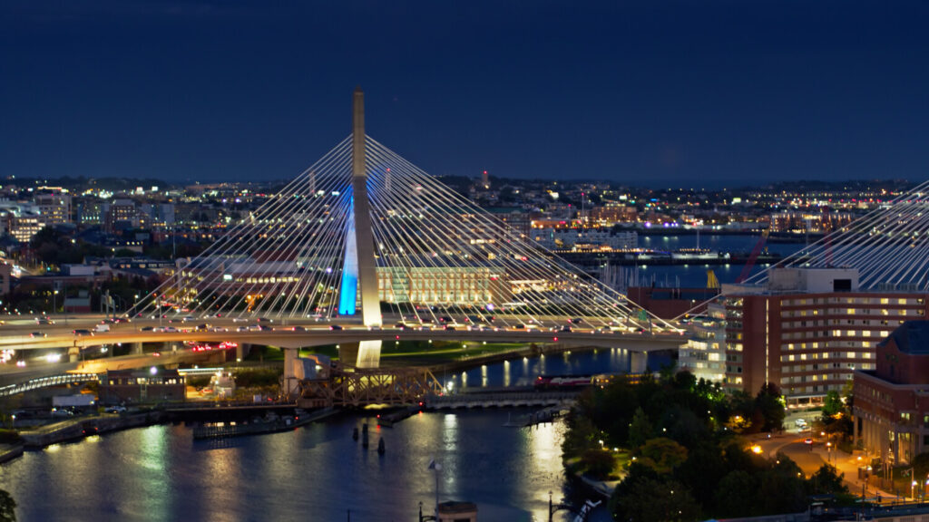 Aerial shot of Boston, Massachusetts at nightfall from over the Charles River, looking towards the Leonard P. Zakim Bunker Hill Memorial Bridge with Boston Harbor beyond. 
